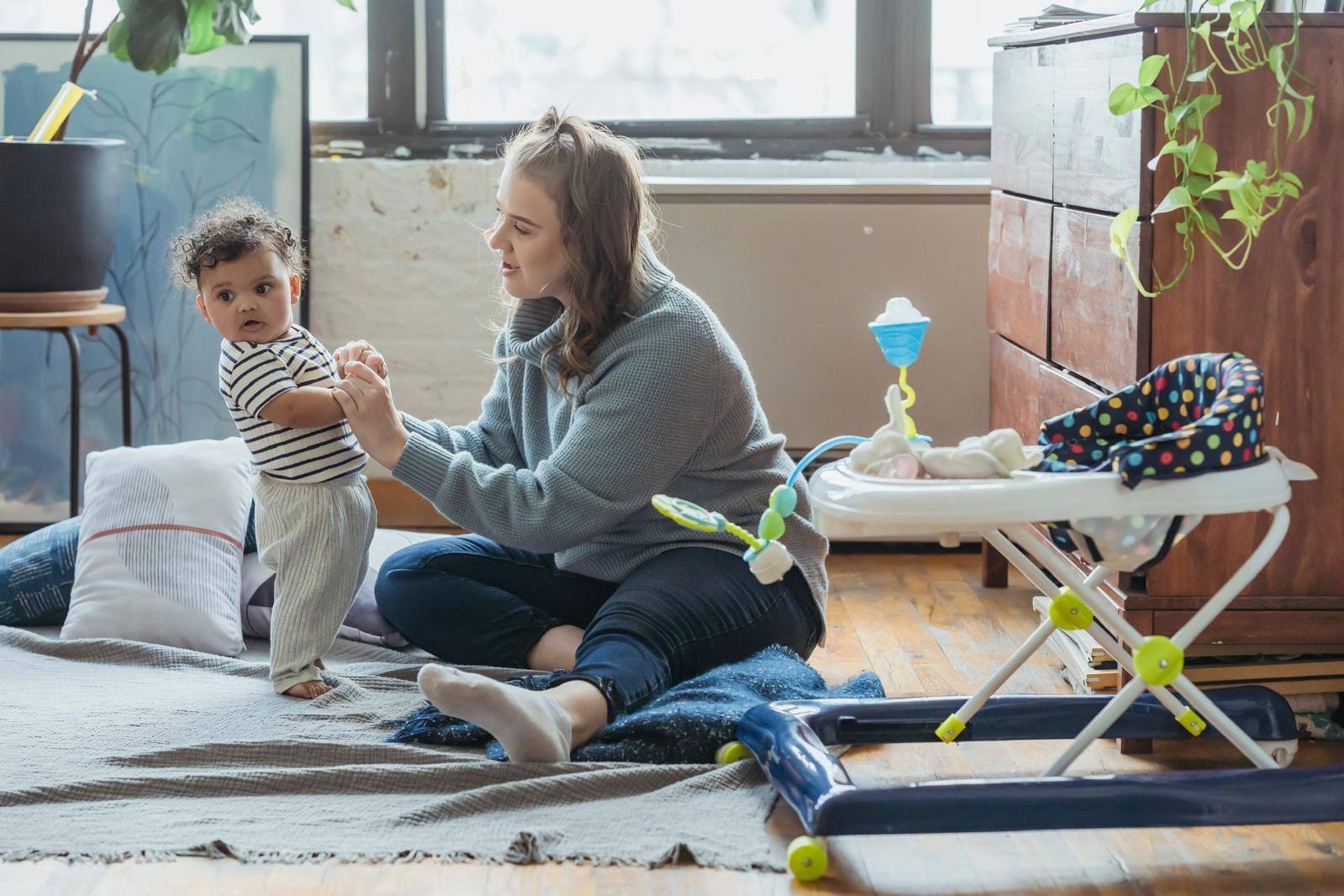 A nanny sitting on the floor helping a baby practise standing