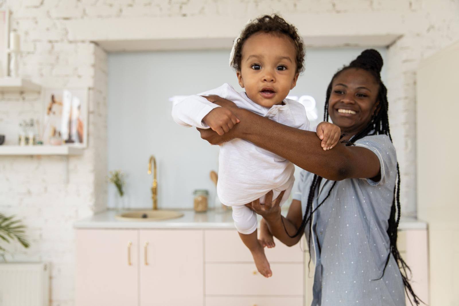 A carer lifting a smiling baby in a bright family kitchen